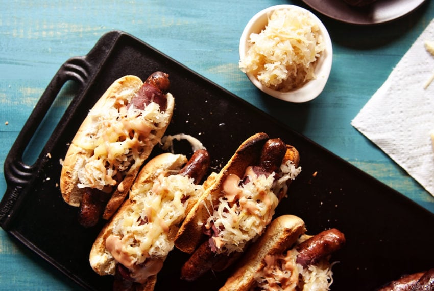 Reuben Hot Dogs on a cast iron griddle topped with sauerkraut, pastrami, thousand island dressing and cheese. More sauerkraut on the side. 