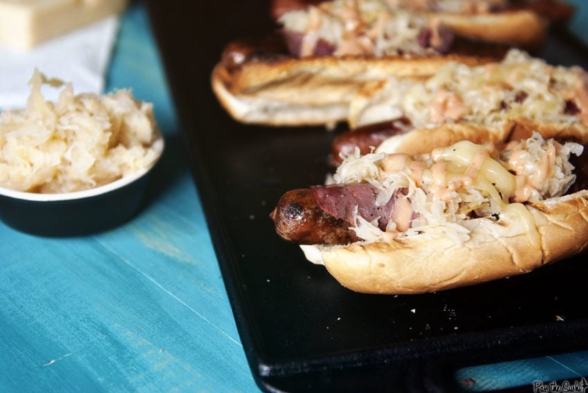 Several grilled rueben hot dogs lined up and topped with kraut, pastrami, cheese and thousand island dressing