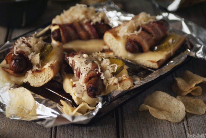 Bacon Wrapped Beer Brats on toasted bread with pickles and onion on aluminium foil and a side of chips