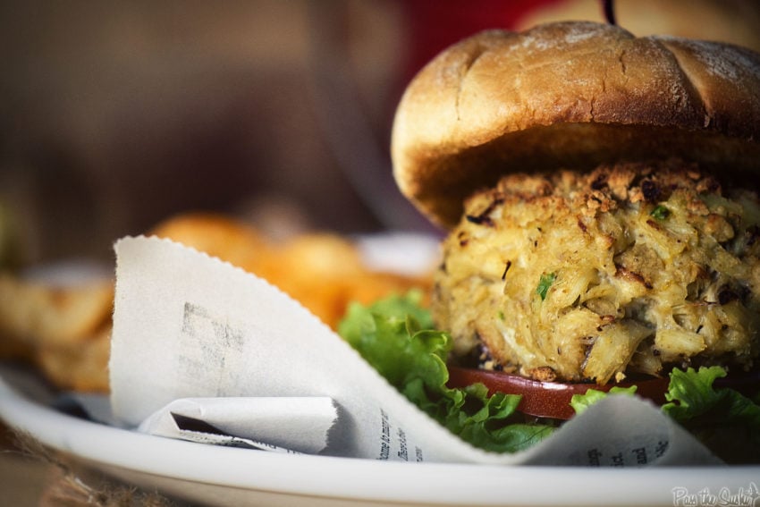 Side shot of a crab cake sandwich, a thick maryland crab cake atop lettuce and tomato. 