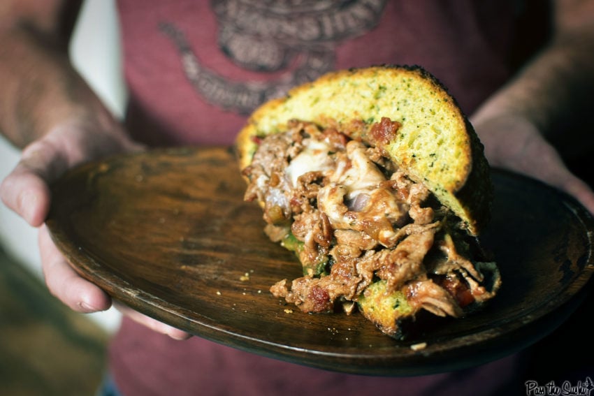 Hands holding a wooden plate with a giant cheesy steak sandwich in the middle. 