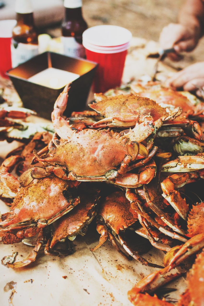 Pile of blue crabs that have been steamed with Old Bay on a paper lined table with hands in the background picking crab. 