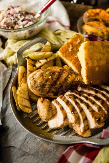 A grilled chicken breast sliced on a metal platter, accompanied by apple slices and cornbread pieces.