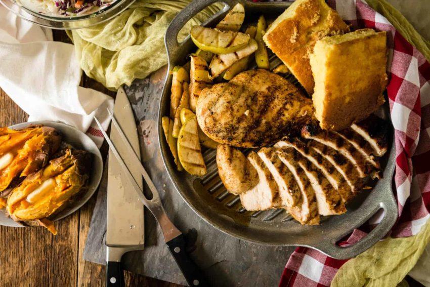 Grilled chicken breast slices, cornbread, and grilled vegetables on a tray with a knife and fork beside it.