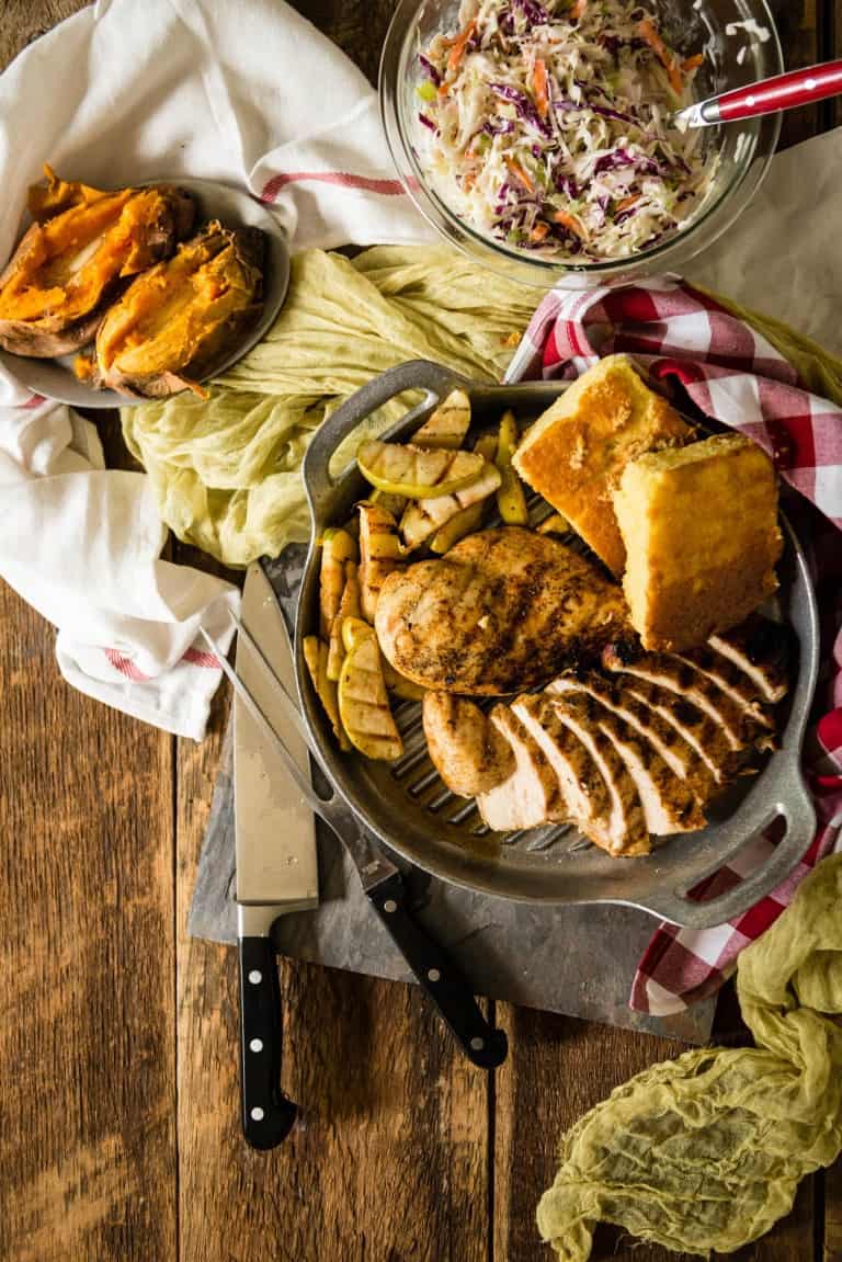 Grilled chicken, cornbread, and roasted vegetables on a tray with a side of coleslaw and baked sweet potatoes.