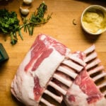 2 racks of lamb, frenched on a cutting board ith herbs, garlic mustard and a knife