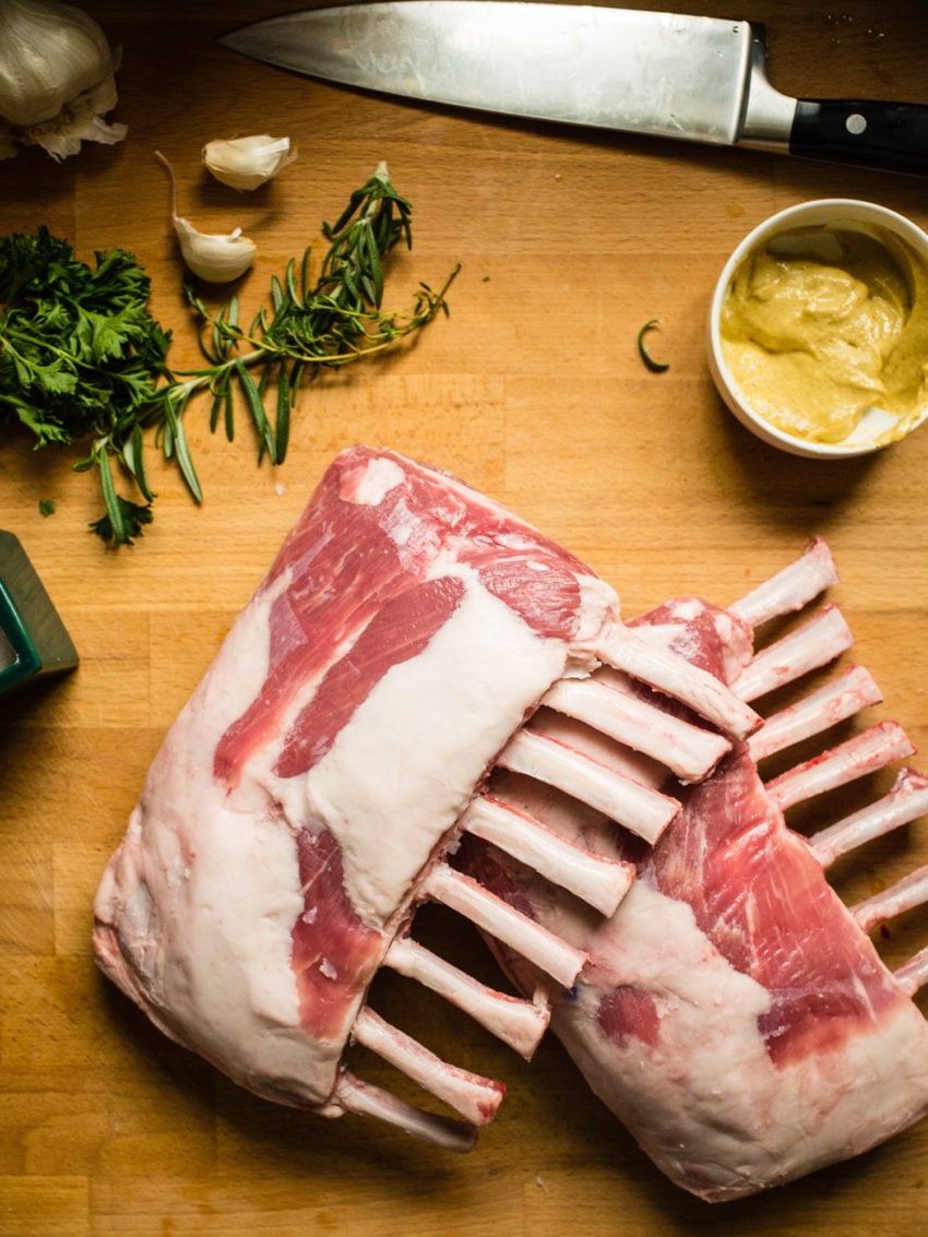 2 racks of lamb, frenched on a cutting board ith herbs, garlic mustard and a knife.