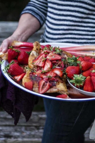 A woman holding a plate of strawberries and rhubarb alongside cast iron roasted chicken.