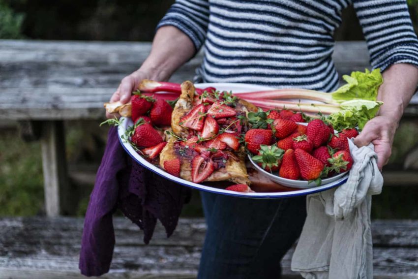 A woman holding a plate of strawberries and rhubarb alongside cast iron roasted chicken.