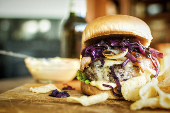 Side view of an irish brat burger with layers of bun, patty, cheese, braised cabbage and onions and a creamy sauce all topped with a bun and chips for nibbling