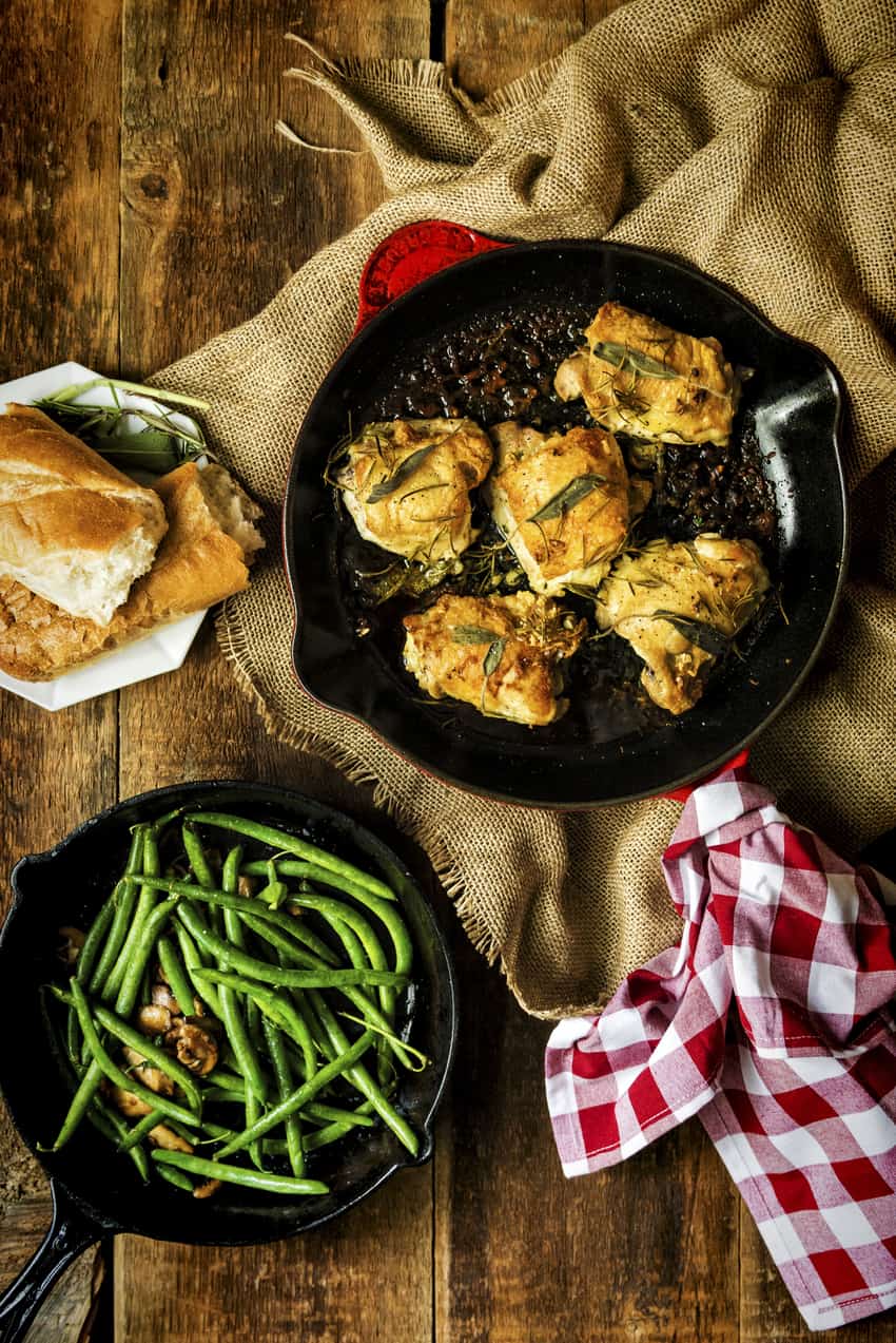 pan seared chicken thighs with crispy skin in red pan with herbs, green beans and bread on the side