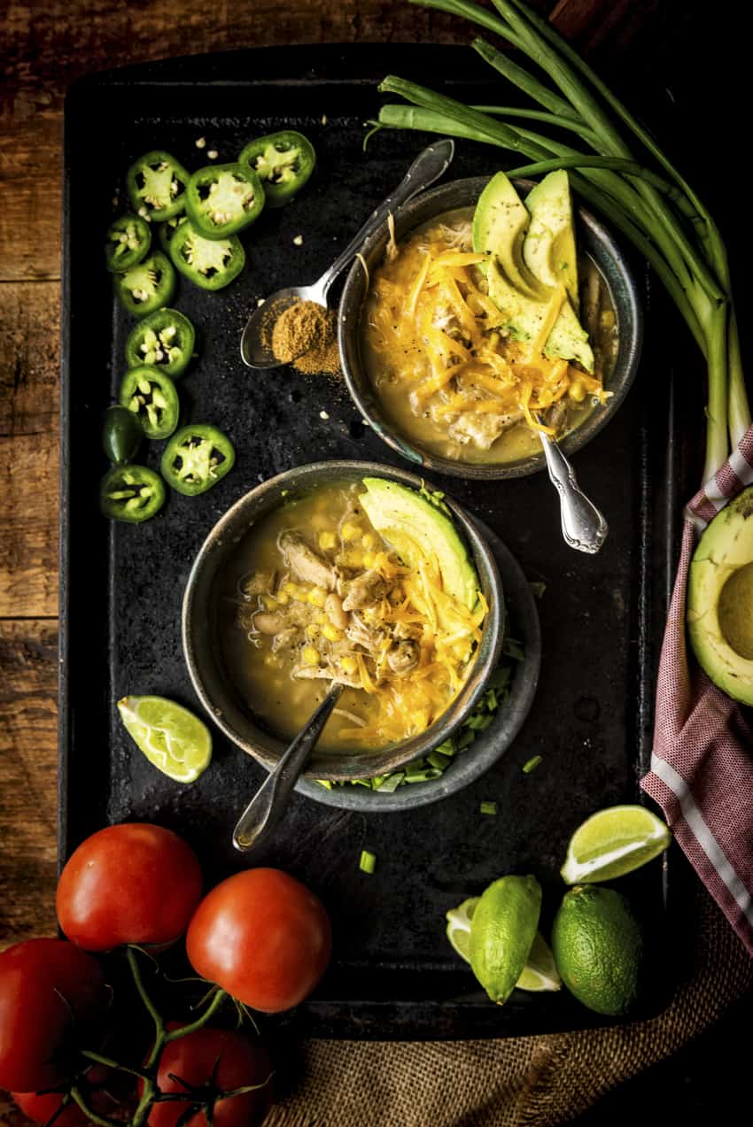 Two bowls of White Chicken Chili topped with avocados and cheese. Extra tomatoes, jalapeños and limes abound.