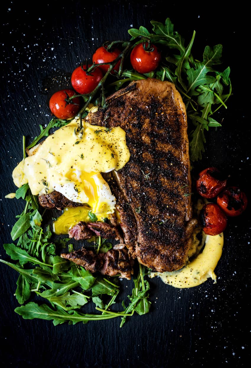 A grilled T-Bone steak with eggs and tomatoes on a plate.