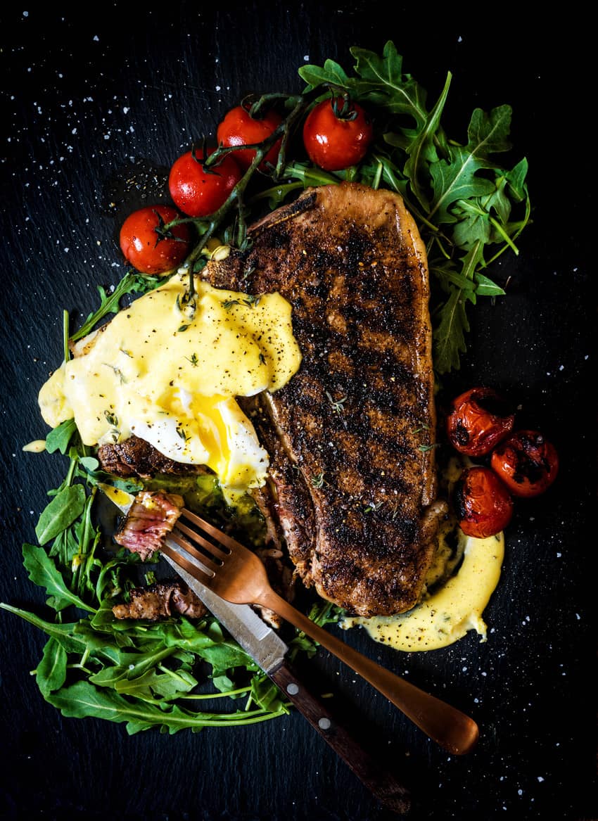 Big t-bone steak with grill marks plated with a broken poached egg atop and bites taken from the tenderloin side.