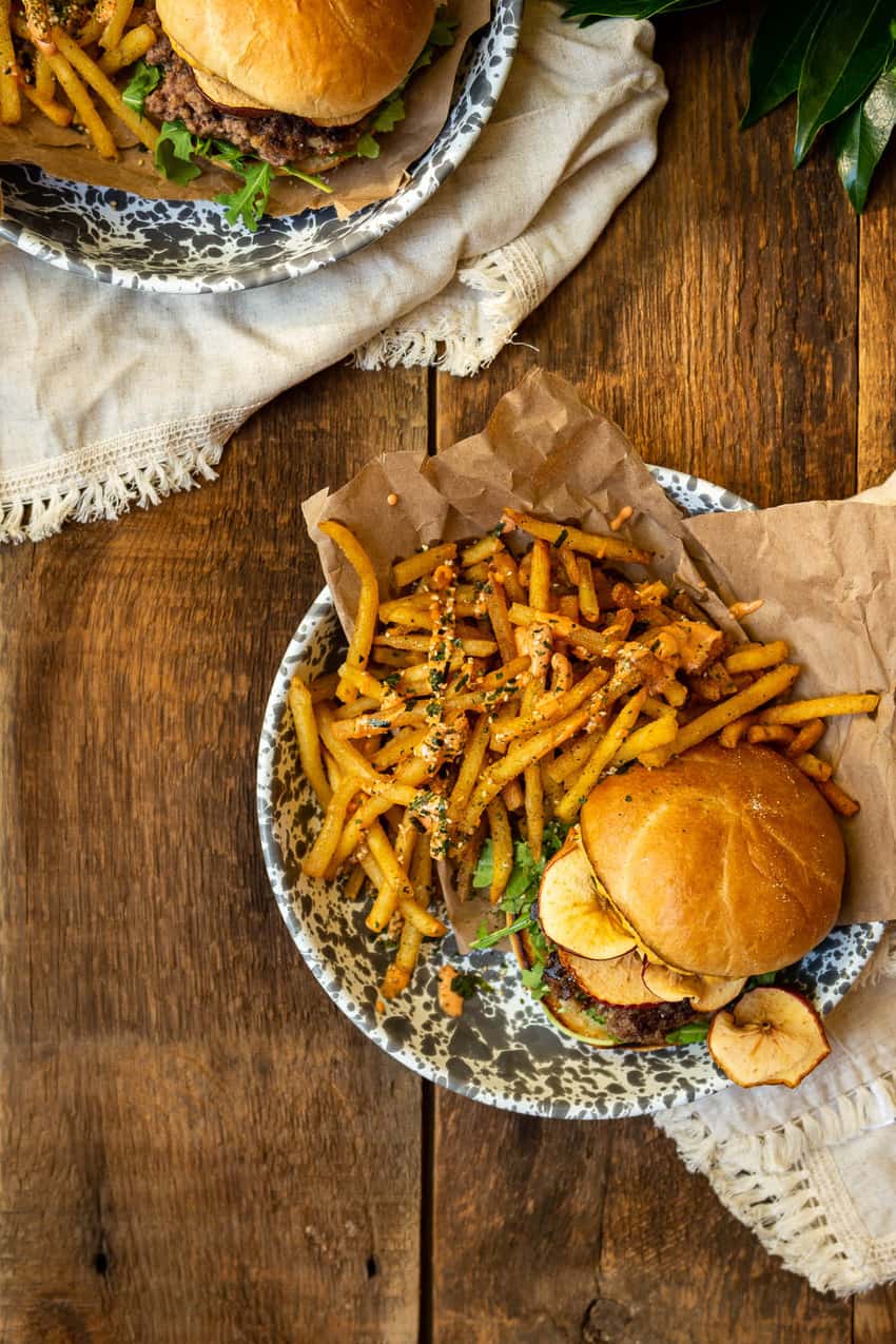 Two plates packed with furikake fries, burgers, apple and fixings. All laying over crinkled brown paper. Dig in!