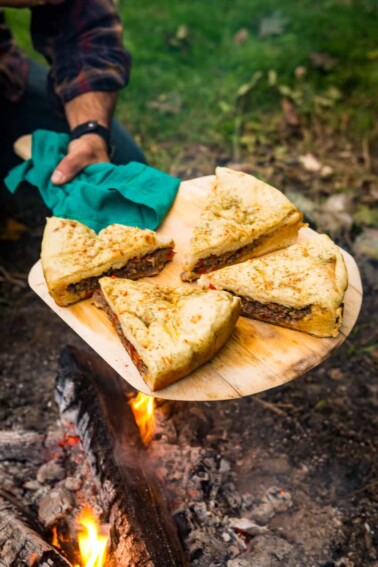 A man is preparing a lamb pizza on a wooden board near a campfire.