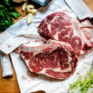 Two raw bone in ribeye steaks with salt, garlic and herbs around them