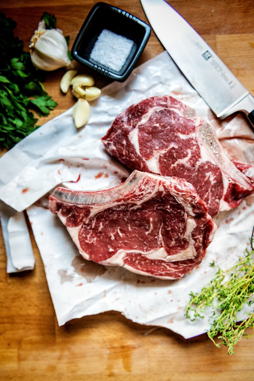 Two raw bone in ribeye steaks with salt, garlic and herbs around them.