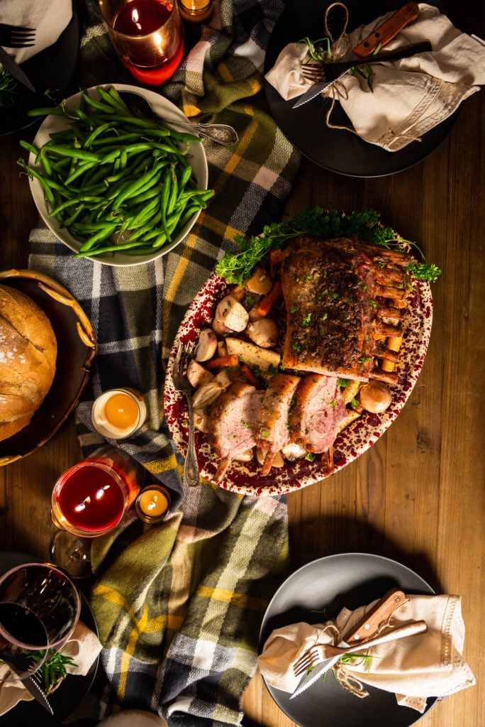 A Rack of Kurobuta Pork served atop a pile of root veggies. Green beans, fresh bread, and holiday place settings fill out this amazing meal.