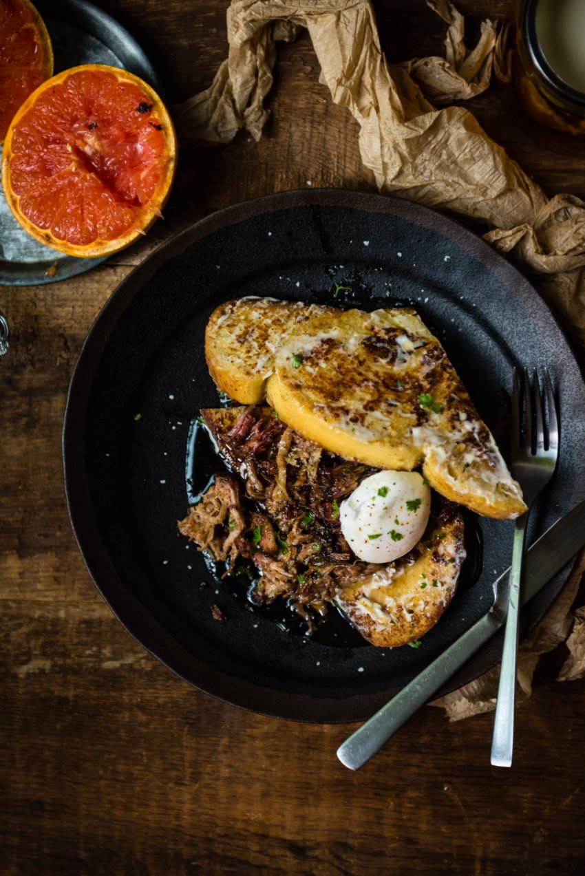 A black plate with two slices of toast, poached egg, shredded pork, and garnished with herbs, placed on a wooden table next to a halved grapefruit.