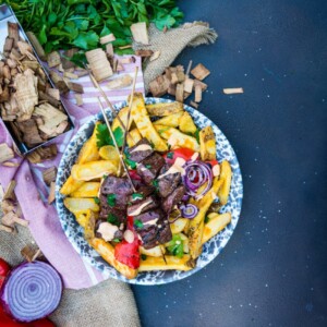 Grilled beef heart kabob recipe over thick cut potatoes with a simple sauce and grilled onions and peppers on a simple background with wood chips and herbs