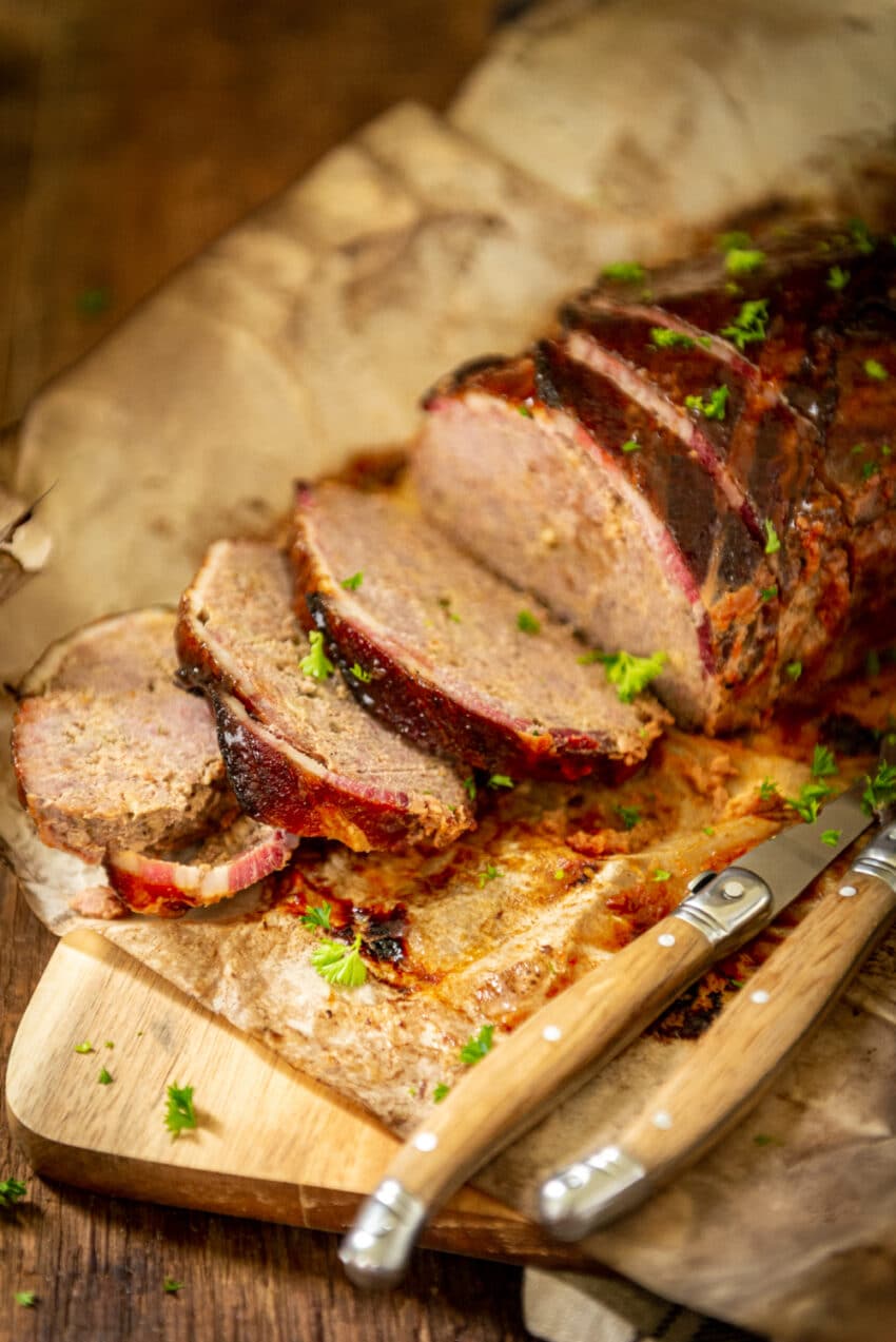 Bacon wrapped smoked meatloaf sliced on a cutting board, garnished with parsley and knife in the foreground. 