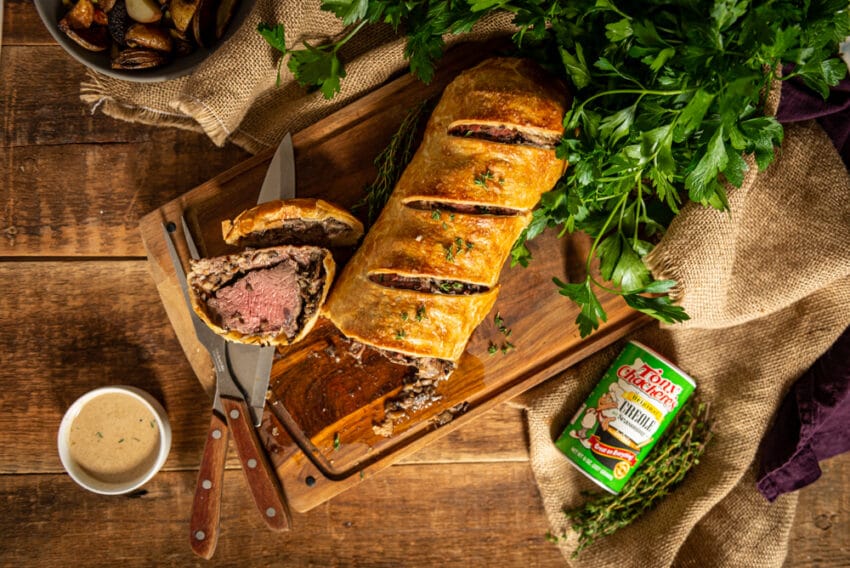 Sliced Beef Wellington arranged on a cutting board with brandy cream sauce and Tony Chachere's spices. 