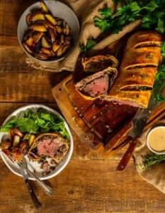 classic beef wellington sliced on a cutting board and a serving on a plate with potatoes and greens.
