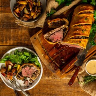 classic beef wellington sliced on a cutting board and a serving on a plate with potatoes and greens.