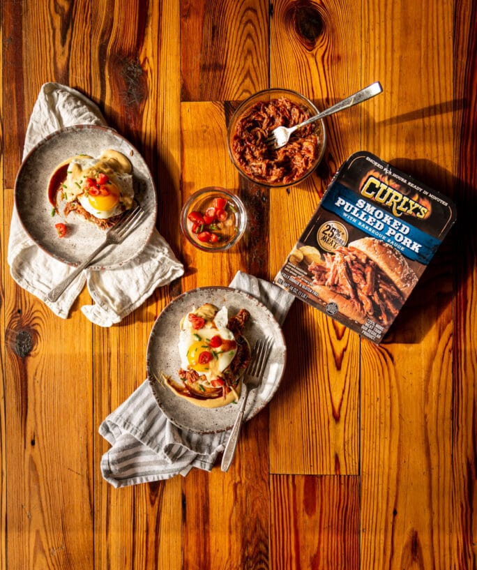 Overhead shot of 2 plates filled with bbq eggs benedict and a package of pulled pork