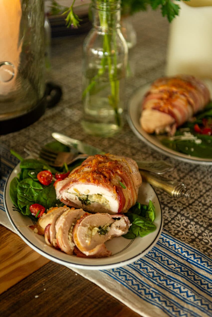 spinach and artichoke stuffed chicken wrapped in bacon, slice on a single dish with salad greens and utensils. 