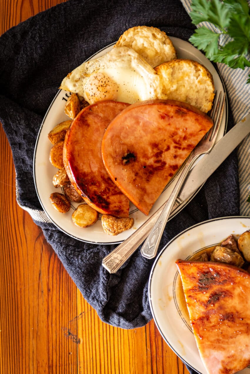 Maple glazed ham steaks sliced and served with red eye gravy over biscuits, egg and potatoes on individual plates