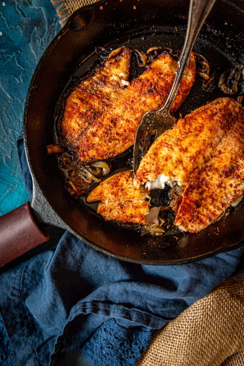 2 tilapia fillets in a cast iron skillet, one showing flakey tender white meat