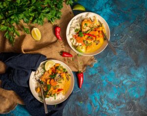 2 bowls of salmon curry over rice with peppers and herbs laying about