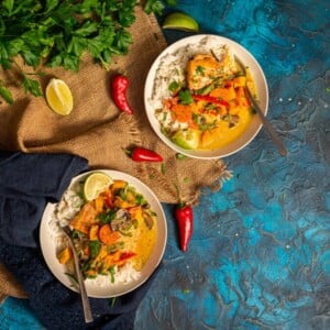 2 bowls of salmon curry over rice with peppers and herbs laying about