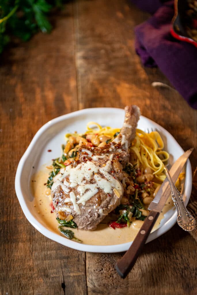 Bone in pork chop served over pasta with a creamy pan sauce and sprinkled with cheese and garnish on plate