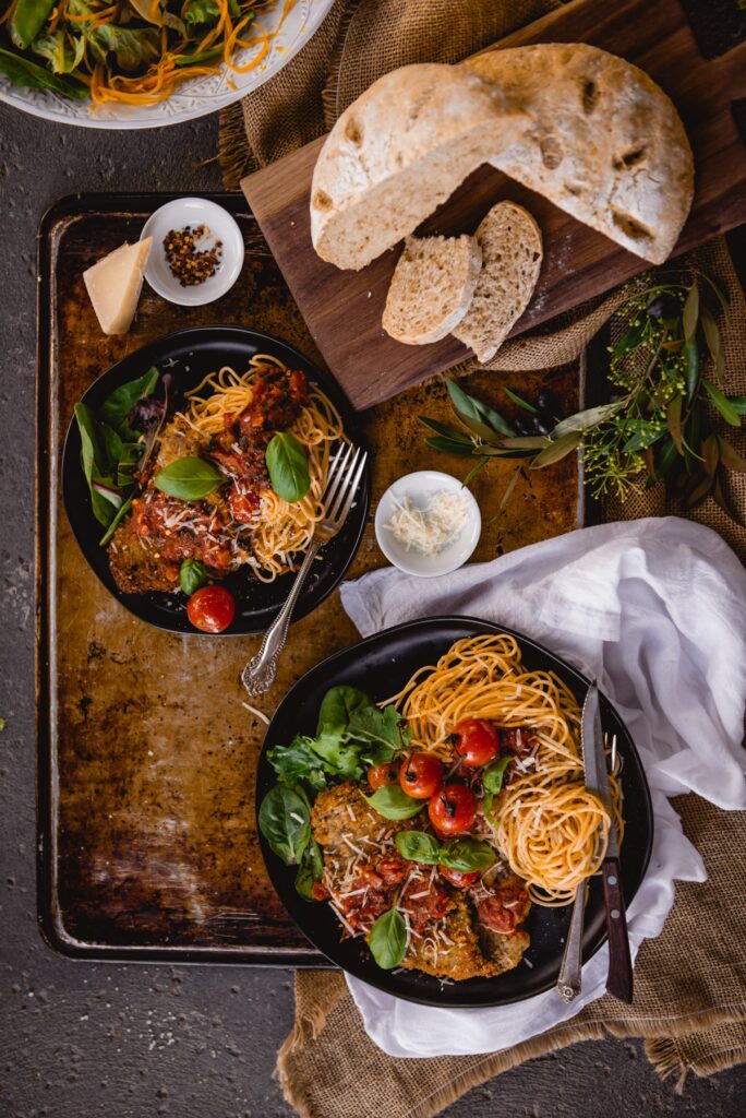 Beef parmesan with pasta on platters with greens and rustic bread