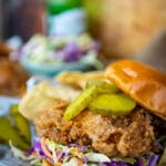 Close up of a fried chicken sandwich on a bun with creamy slaw and pickles on top with drink and more chicken in the background