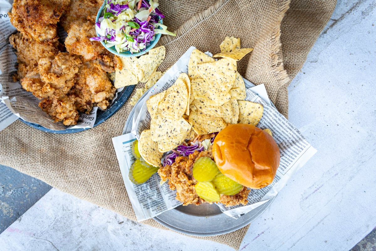 Above shot of fried chicken sandwich with chips and a platter of extra fried chicken and slaw