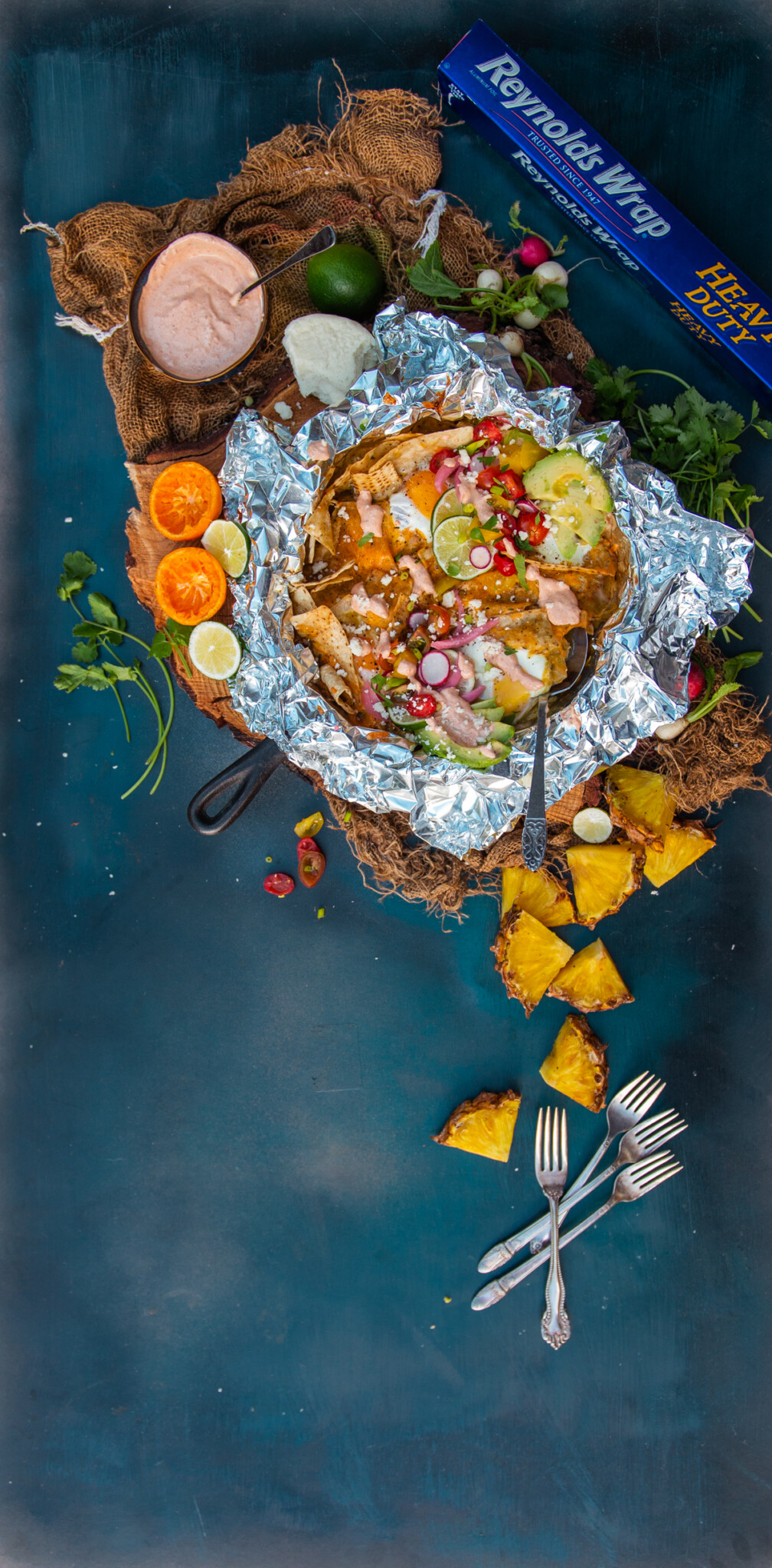 above shot of finished chilaquiles recipe in cast iron with foil for campfire or gas grill. pineapples and other topping scattered around on a blue background