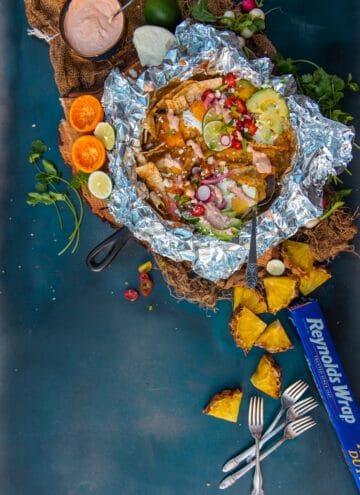 above shot of finished chilaquiles recipe in cast iron with foil for campfire or gas grill. pineapples and other topping scattered around on a blue background