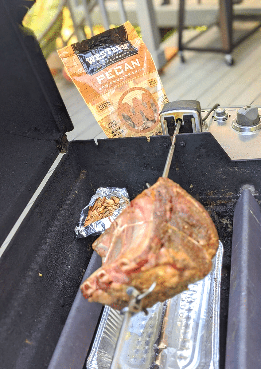 Standing rib roast on rotisserie with pecan wood foil packet on gas grill. 