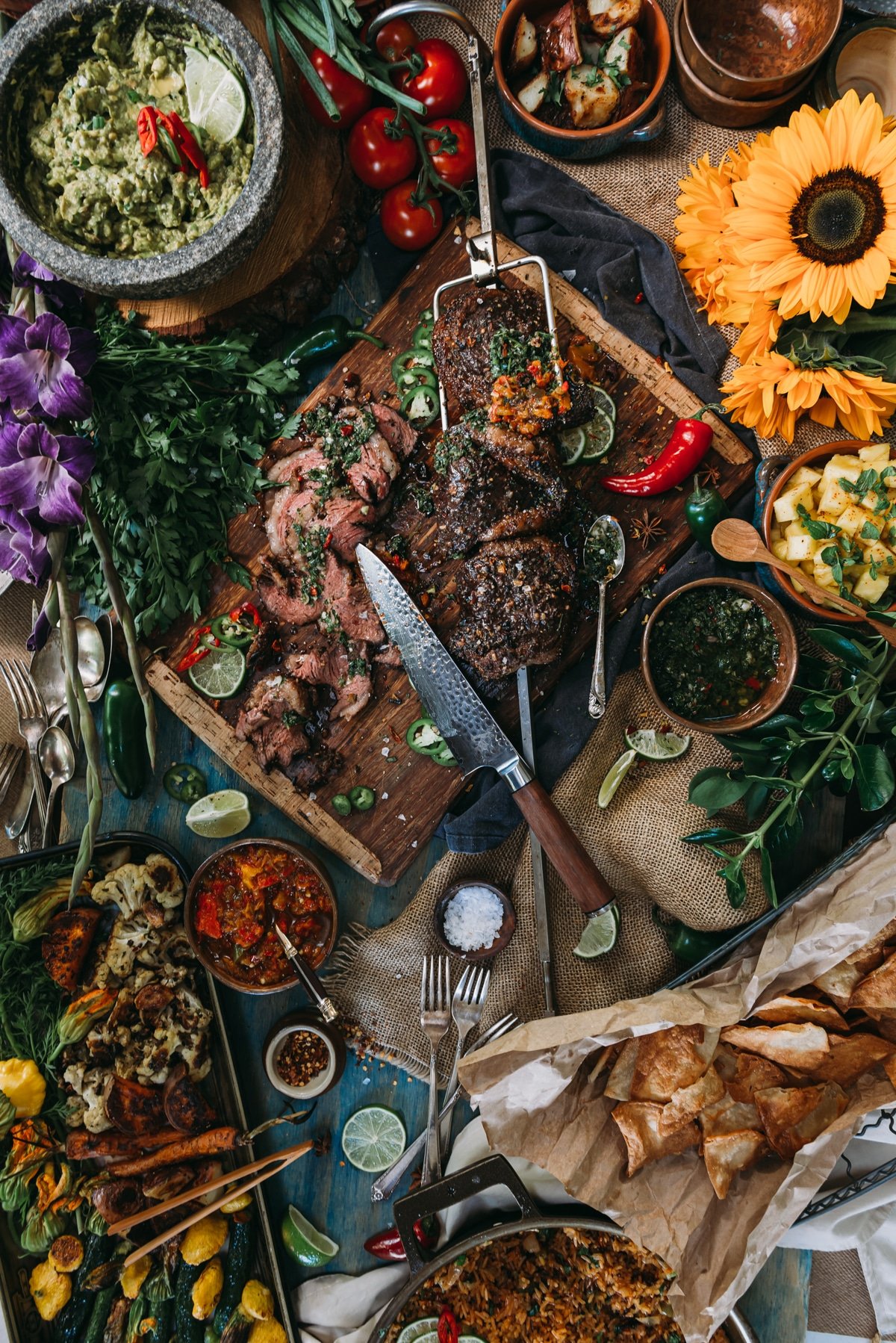 Gas grilled picanha being sliced - shown from above on a table filled with guacamole, flowers, peppers, tortilla chips and more. 