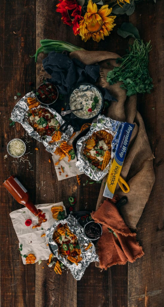 above shot of messy waffle fries in foil packets covered in creamy jalapeno popper dip with various other ingredients laying about 
