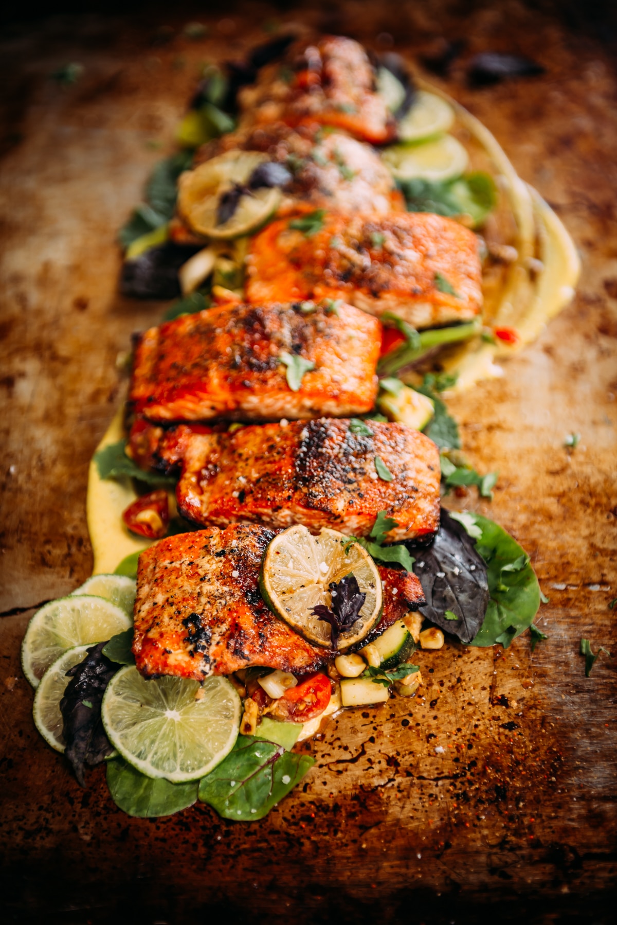 long shot of a salmon filet that has been portioned and grilled plated over greens with lime slices and thai basil as garnish.