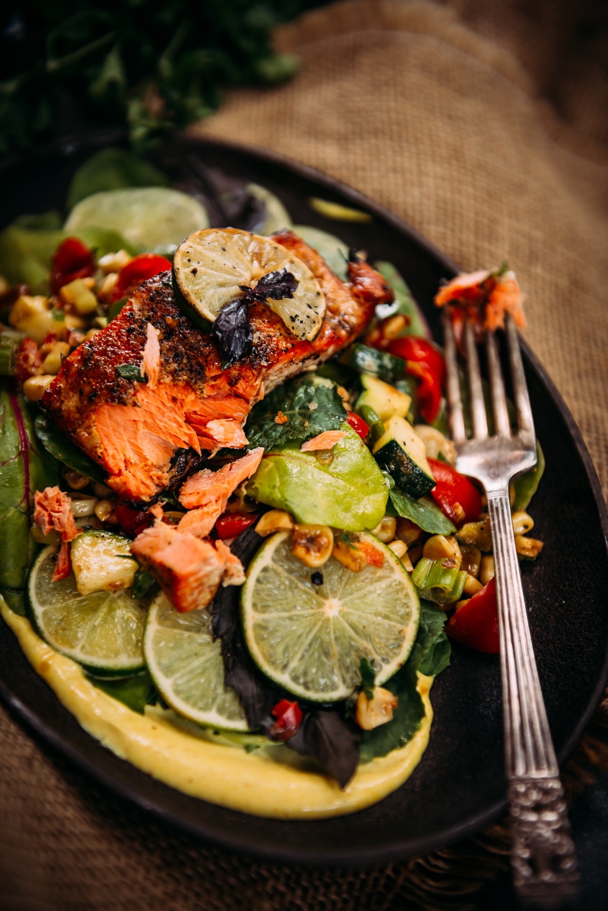 one salmon portion, grilled with a fork taking a section out to show inner flakiness over succotash, salad, and cardamom aioli.