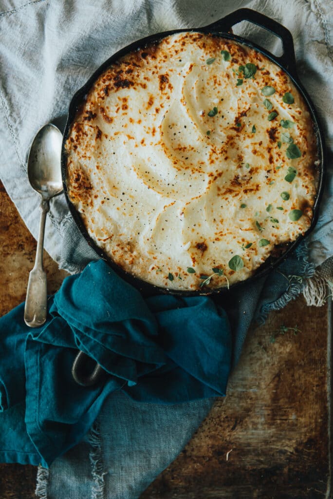 creamy mashed potatoes on top of a hearty minced beef shepherd's pie in a cast iron skillet over blue cloths