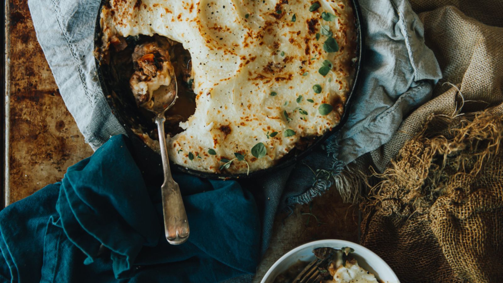 A partially eaten dish of shepherd's pie, crafted from a treasured recipe, sits with a serving spoon on a textured cloth, surrounded by rustic fabric. A small bowl of pie with a fork waits nearby, inviting another taste.