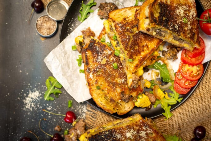 close up of an epic grilled cheese sandiwch, using leftover cheeseburgers, scrambled eggs and cheese on a plate with sliced tomatoes and arugula