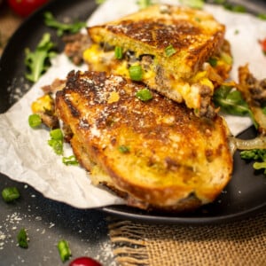 close up of grilled cheese, filled with breakfast eggs, leftover burger, and loads of cheese with various food garnish around on black plate